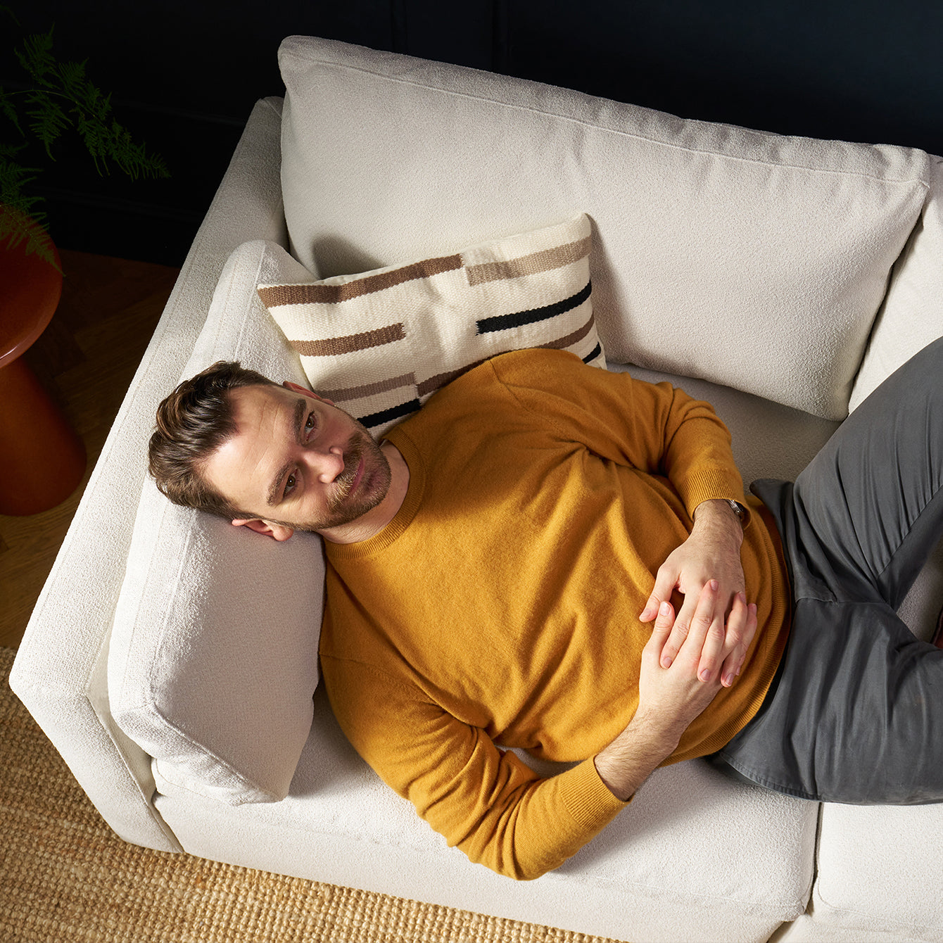 A photo from above of a man in a yellow jumper and grey trousers, laying on a modern white textured sofa. He is resting his head on the arm lumbar cushion and relaxing.
