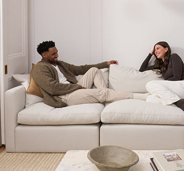 A front on image of a male and female couple, sitting on a white feather filled modern modular sofa. They are relaxing with their legs up on the sofa and looking at each other.